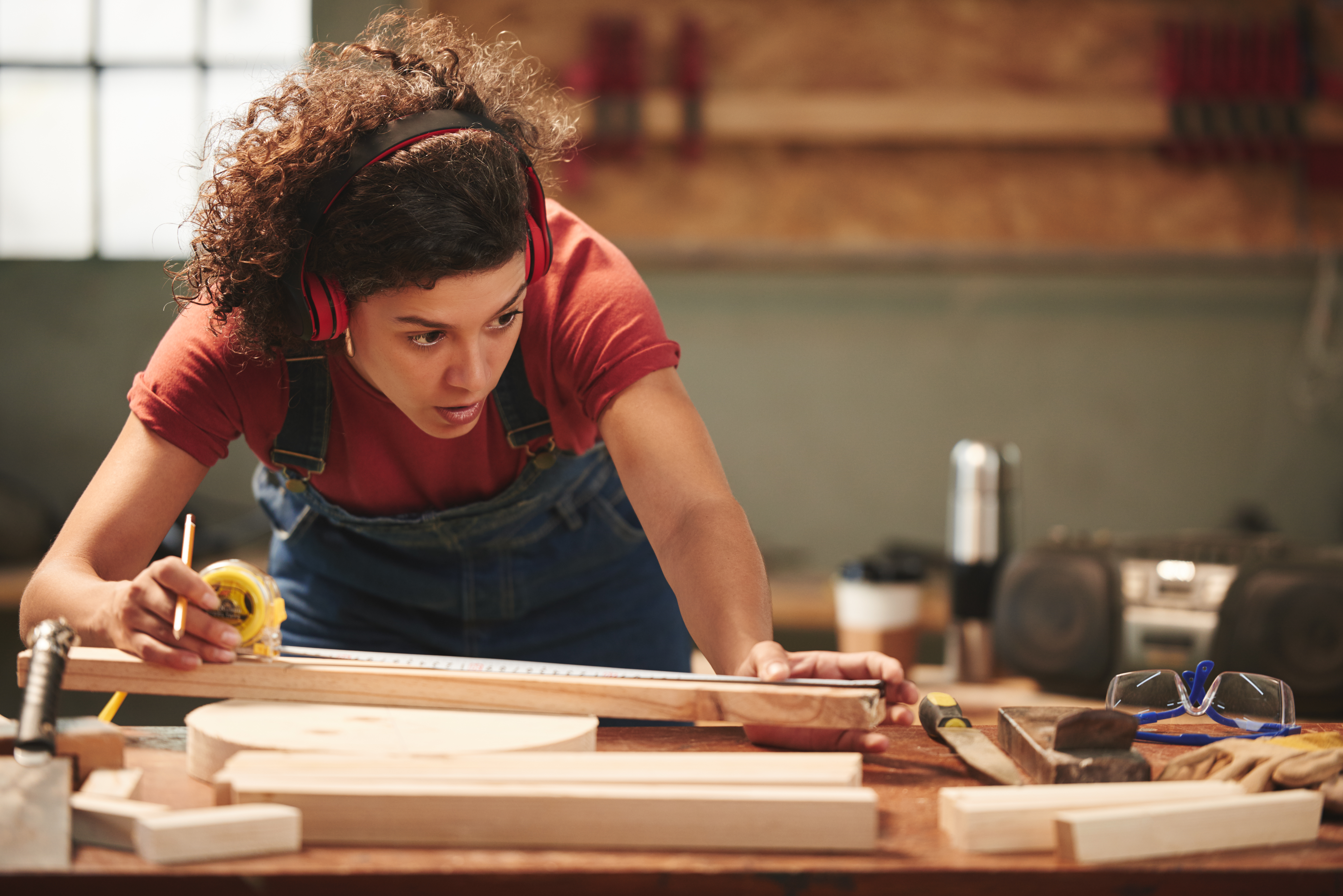 Young concentrated curly woman in ear defenders and overall measuring wooden plank with tape measure