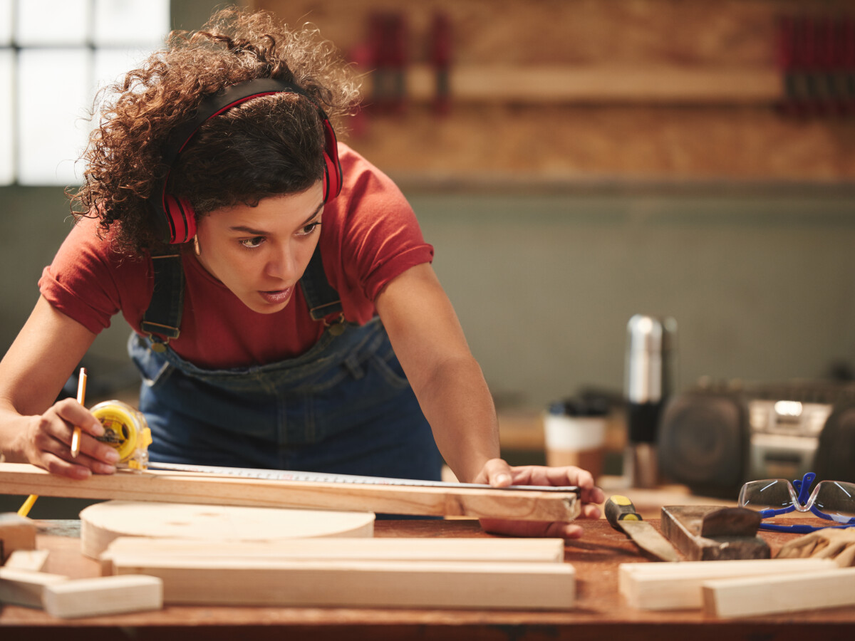 Young concentrated curly woman in ear defenders and overall measuring wooden plank with tape measure